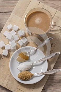 Lump sugar, cane sugar, loose white sugar and buckwheat honey on a teaspoon lie next to a glass of coffee with milk.