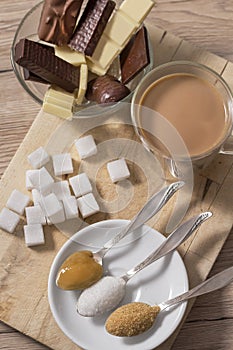 Lump sugar, cane sugar, loose white sugar and buckwheat honey on a teaspoon lies next to a glass with coffee and milk. In the back