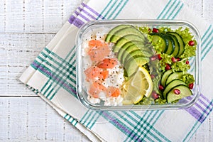 Lunch box: rice, salmon, salad with cucumber, avocado, greens on a white wooden background. Fitness food.