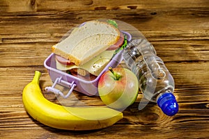 Lunch box with sandwiches, bottle of water, banana and apple on wooden table