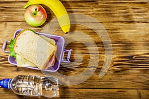 Lunch box with sandwiches, bottle of water, banana and apple on wooden table. Top view, copy space