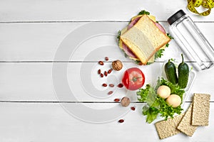 Lunch boxes with sandwich and fresh vegetables, bottle of water, nuts and eggs on white wooden background. Top view with