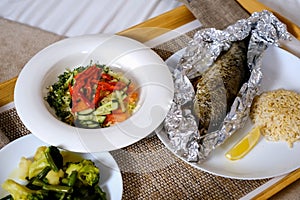 Lunch with fish, salad, rice and boiled vegetables on wooden tray in hotel room.