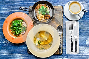 Lunch: frying pan with meat and herbs, yellow soup, salad of vegetables and herbs, coffee and cutlery, on a blue denim background.