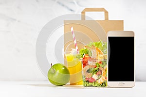 Lunch set with seafood salad with shrimps, orange juice, green apple, packet, blank phone in white interior. Mockup.