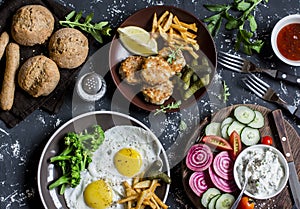 Lunch table - fried eggs, fish balls, potato chips, vegetables, sauces, homemade bread on a dark background.