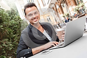 Lunch time. Young man in suit sitting at cafe on city street drinking hot coffee browsing internet on laptop looking