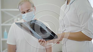 Lungs x-ray in female hands with unrecognizable doctor explaining roentgen to mid-adult man in face mask. Professional