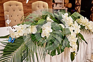 Lush floral arrangement of orchids and monstera leaves on wedding table. Wedding presidium in restaurant, copy space.