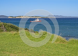 Lush green grass sea shore, deep blue sea with red fisherman boat and steep cliffs, north east coast of Corfu in Greece