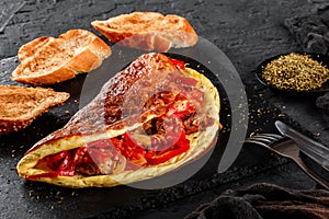 Lush omelet with meat, vegetables, cheese and herbs on black slate over dark background. Healthy breakfast. Top view, flat lay