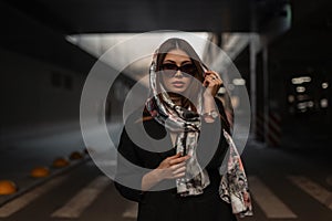 Luxurious attractive young woman with sexy lips with stylish silk scarf on head in trendy black coat in fashionable sunglasses