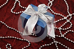 Luxurious blue gift box with a silver ribbon on a red shiny cloth and decorative silver string of pearls.