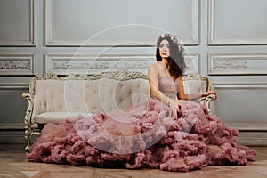 Luxurious girl in an evening cloudy dress sitting on a sofa in the studio interior.