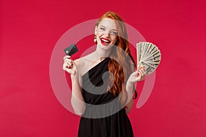 Luxury, beauty and money concept. Elegant good-looking redhead woman being rich, wear luxurious black dress, holding