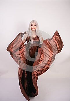 Luxury blonde caucasian model girl in chocolate color long evening dress made of pleated fabric waving a flying dress and stands o