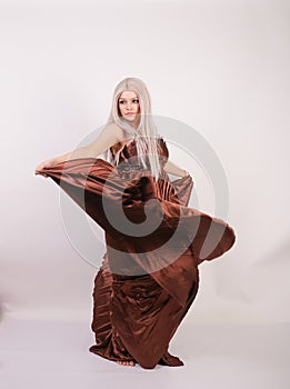 Luxury blonde caucasian model girl in chocolate color long evening dress made of pleated fabric waving a flying dress and stands o