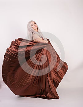 Luxury blonde caucasian model girl in chocolate color long evening dress made of pleated fabric waving a flying dress and stands o