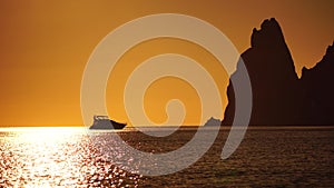 Luxury cruise trip. Red burning sunset over the sea with silhouette of rich yacht and rocky volcanic cliff. Abstract