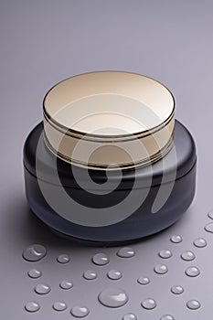 Luxury face cream jar with golden lid and matte glass on grey background with waterdrops. Packaging presentation, unbranded posh