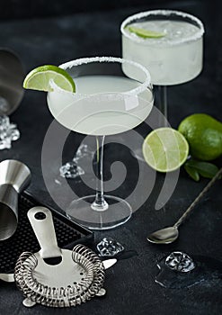 Luxury glasses of Margarita cocktail with fresh limes and bar mat with strainer and jigger, with spoon and ice cubes on black