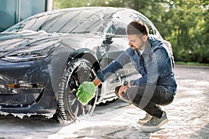 Luxury modern electric car in soap foam outdoors at car wash service. Side view of handsome young Caucasian man using