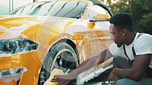 Luxury modern sport car in soap foam outdoors at car wash service. Side view of handsome young African man using yellow