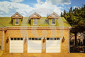 Luxury three car garage with carriage lights between doors set in brick house with green wood shingle roof and dormers