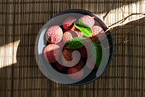 Lychees in black bowl over brown bamboo mat in hard light.