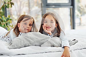 Lying down on bed and looking at the camera. Two cute little girls indoors at home together. Children having fun