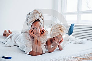 Lying down on white bed together. Young mother with her daugher have beauty day indoors in room