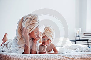 Lying down on white bed together. Young mother with her daugher have beauty day indoors in room