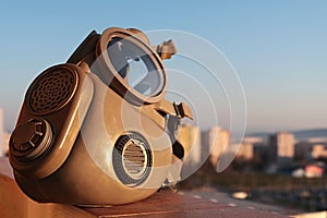 M10 Military grade gas mask with inhale and exhale valves visible, sunbathing in morning sunshine, cityscape in background.