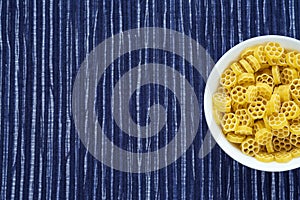 Macaroni ruote pasta in a white bowl on a striped white blue cloth background with a side. Close-up with the top. Free