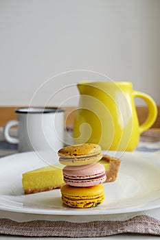 Macaroons and lemon tart on white plate. Whine enamel mug, yellow ceramic jug. Still life in pastoral style aesthetics.