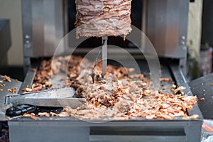 Machine for cooking shawarma, meat layers strung on a skewer, chopped meat is on grill surface, tongs and knife, front view