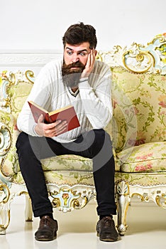 Macho on disgusted face reading book. Scandalous bestseller concept. Man with beard and mustache sits on baroque style