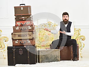 Macho elegant on surprised face sits, confused at end of packing, near pile of vintage suitcases. Man with beard and