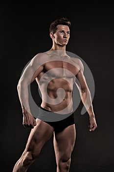 Macho with muscular body posing as bodybuilder, black background. Bodybuilder in black underpants shows his muscles