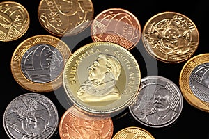 An assortment of Brazilian coins on a black reflective background with a South African one ounce gold Krugerrand coin