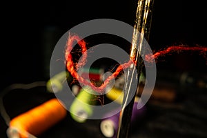 Needle eye threaded with piece of red thread on black background with colored spools of thread. Steel tailor needle with