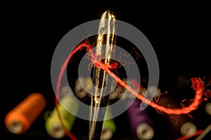 Needle eye threaded with piece of red thread on black background with colored spools of thread. Steel tailor needle with