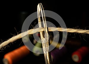 Needle eye threaded with piece of white thread on black background with colored spools of thread. Steel tailor needle