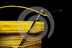 A macro image of a needleâ€™s eye threaded with a piece of yellow thread on a field of solid black.  The needle is entrapped in