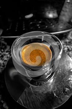 Macro view of a glass cup with freshly brewed black coffee with golden foam with small bubbles in a metal coffee saucer on top of