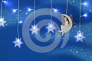 Magical christmas image of little white fairy with glitter wings sitting on the moon over blue background and silver snowflake gar