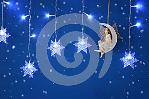 Magical christmas image of little white fairy with glitter wings sitting on the moon over blue background and silver snowflake gar