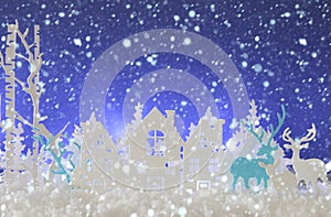 Magical Christmas paper cut winter background landscape with houses, trees, deer and snow in front of blue background.