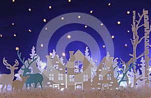 Magical Christmas paper cut winter background landscape with houses, trees, deer and snow in front of night starry sky background.