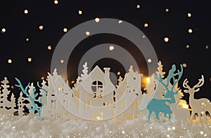 Magical Christmas paper cut winter background landscape with houses, trees, deer and snow in front of night starry sky background.